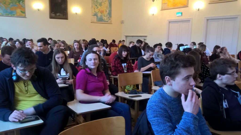 Młodzież słucha wykładu siedząc w ławkach na sali wykładowej
