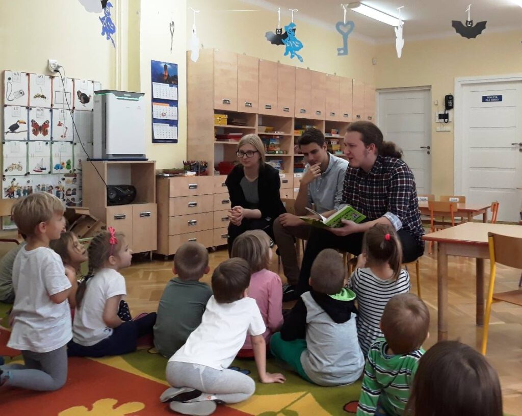 Troje słuchaczy czyta książkę grupie siedzących na podłodze przedszkolaków