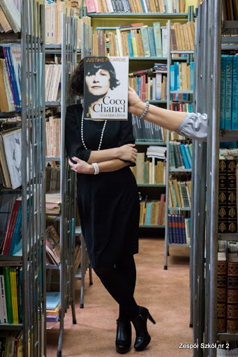 Kobieta stojąca pomiędzy regałami bibliotecznymi z twarzą zasłonietą okładką książki