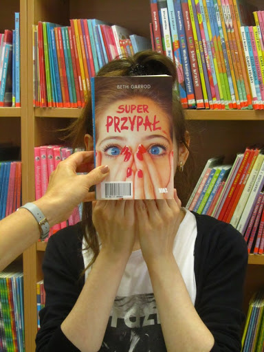 Kobieta na tle regału z książkami zasłaniająca twarz książką