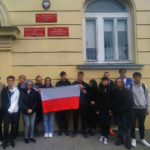 Kilkoro uczniów stoi przed budynkiem szkoły i trzyma rozłożoną polską flagę