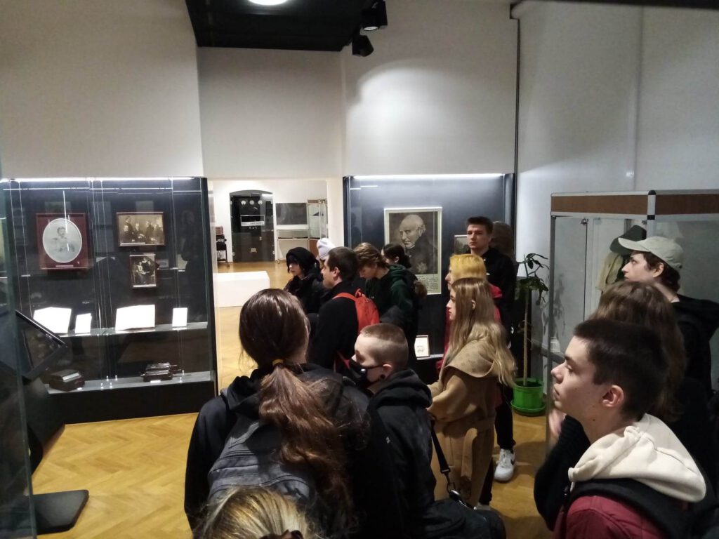 Młodzi ludzie oglądają wystawę muzealną. stoją przed gablotami i słuchają przewodniczki