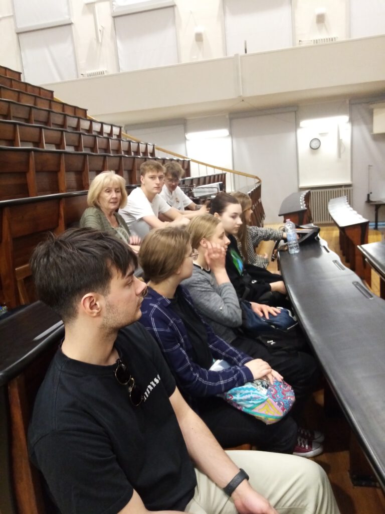 Grupka młodzieży i nauczycielka siedzą w ławkach sali wykładowej