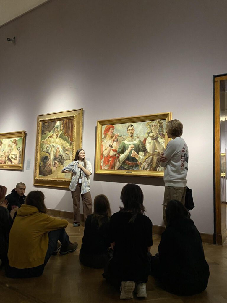 Grupa młodzieży siedzi na podłodze patrząc na wiszący na ścianie obraz i słucha wykładu stojącej przed nim przewodniczki