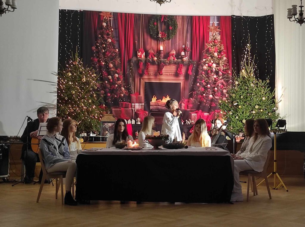 Trzy kobiety siedzą przy stole na tle świątecznej dekoracji. Czwarta kobieta stoi i śpiewa do trzymanego w ręku mikrofonu