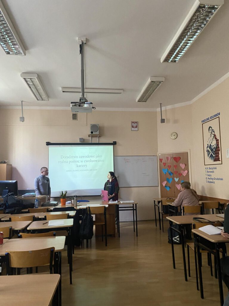 Mężczyzna i kobieta stoją w klasie na tle ekranu. Na ekranie wyświetlana jest prezentacja.