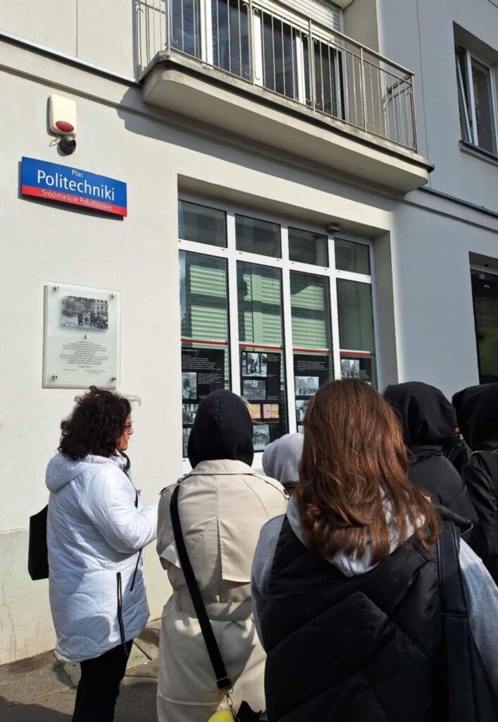 Zdjęcie przedstawia uczniów klasy 2PH3 w towarzystwie opiekuna szkolnego wolontariatu, Pani Iwony Małkowskiej. Grupa znajduje się przy ul. Lwowskiej 2a i patrzy na tablicę pamiątkową.