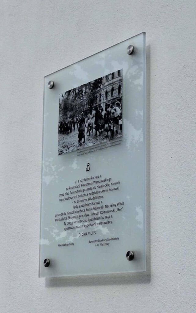 Zdjęcie tablicy pamiątkowej. Na tablicy znajduje się zdjęcie z upadku Powstania Warszawskiego.Tekst na tablicy: "4 i 5 października 1944 r. po kapitulacji Powstania Warszawskiego przez plac Politechniki przeszła do niemieckiej niewoli część walczących do końca oddziałów Armii Krajowej - tu żołnierze składali broń. Tędy 5 października 1944 r. poszedł do niewoli dowódca Armii Krajowej i Naczelny Wódz Polskich Sił Zbrojnych gen. dyw. Tadeusz Komorowski "Bór". Tą drogą we wrześniu i październiku 1944 r. opuszczali miasto wypędzani warszawiacy. Gloria victis"