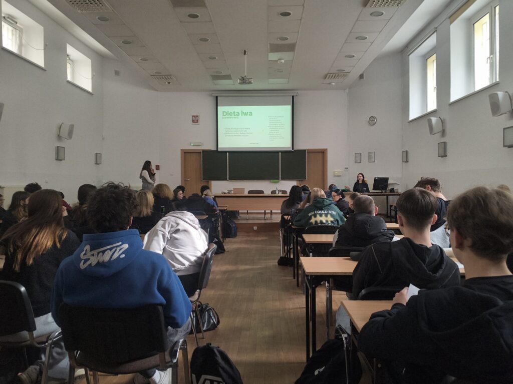 Zdjęcie wykonane z tyłu sali: przedstawia plecy uczniów uczestniczących w warsztatach, na froncie stoją prelegentki. Slajd prezentacji wyświetlany na ekranie mówi o diecie lwa (carnivore diet).