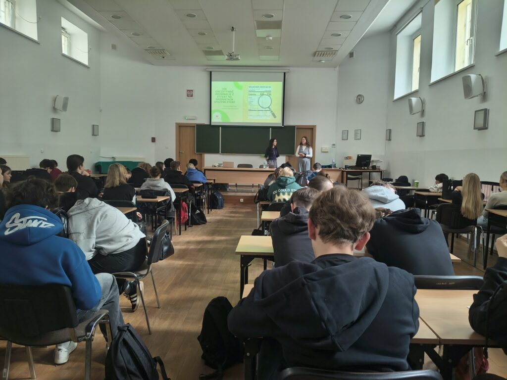 Zdjęcie wykonane z tyłu sali: przedstawia plecy uczniów uczestniczących w warsztatach, na froncie stoją prelegentki. Slajd prezentacji wyświetlany na ekranie zawiera informacje o tym, jak odczytywać informacje z etykiet produktów spożywczych.